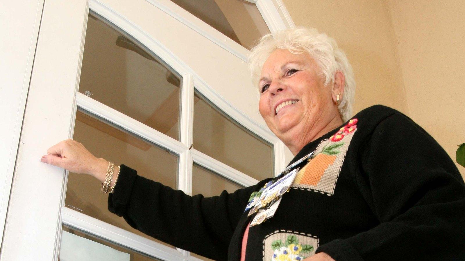 A smiling visiting nurse with a lanyard badge opens a front door for a home visit